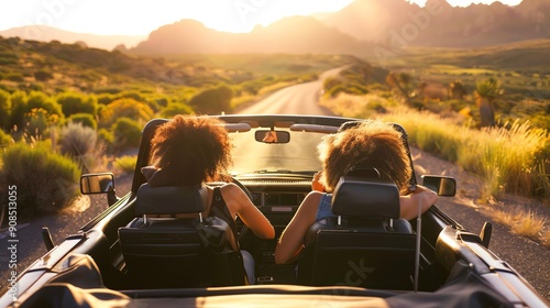 Two women driving a convertible down a road in a mountainous region.