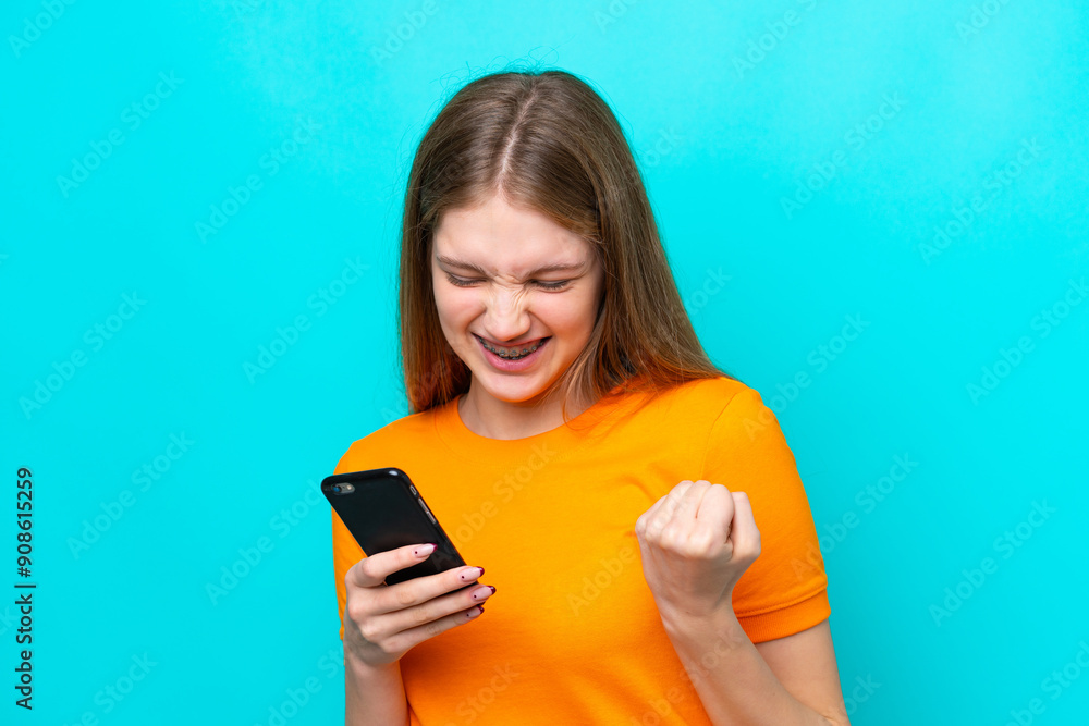 Teenager Russian girl isolated on blue background using mobile phone and doing victory gesture Teenager Russian girl isolated on blue background using mobile phone and doing victory gesture