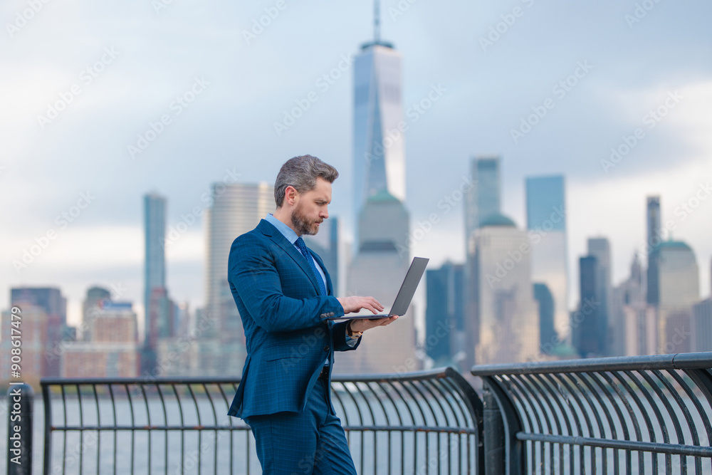 Businessman with laptop on financial district streets of New York City. Business in Manhattan. Business man on cityscape in NYC. Business man in suit outdoor. Office worker. Mature business manager. Businessman with laptop on financial district streets of New York City. Business in Manhattan. Business man on cityscape in NYC. Business man in suit outdoor. Office worker. Mature business manager.