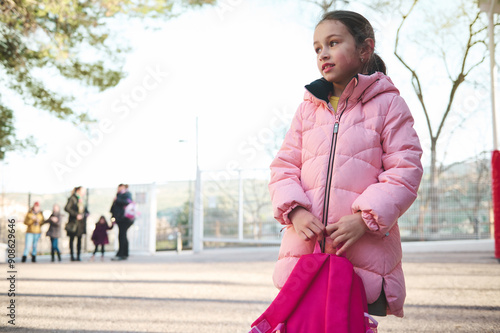 Young girl in pink jacket holding backpack, standing outdoors on a clear day photo