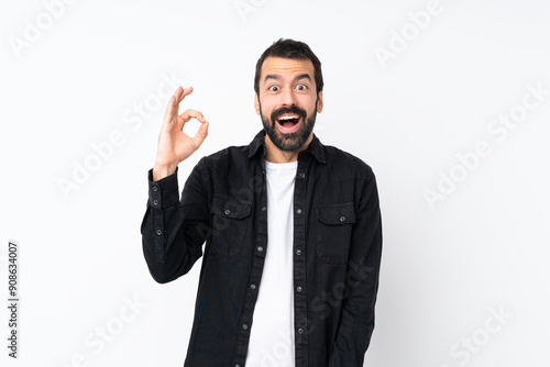 Young man with beard over isolated white background surprised and showing ok sign