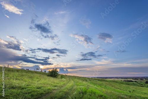 A dirt path meanders through a grassy hillside under a sky filled with fluffy white clouds and a vibrant sunset.