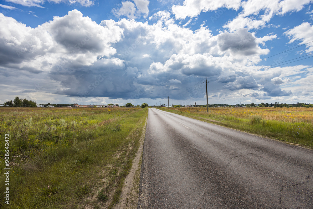 A tranquil rural road leads into the distance, flanked by fields, while dramatic clouds hover overhead in the vibrant afternoon sky. A tranquil rural road leads into the distance, flanked by fields, while dramatic clouds hover overhead in the vibrant afternoon sky.