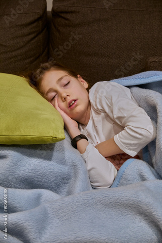 Vertical photo of 10-year-old girl sleeping on the sofa with a green cushion as a pillow and covered with a blue blanket