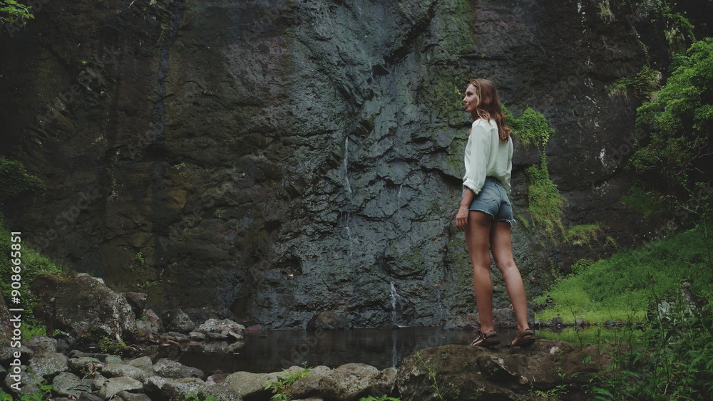 Naklejka premium A woman stands admiring a waterfall in French Polynesia, her back to the camera, as she gazes at the cascading water and lush greenery.