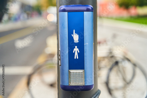 A pedestrian signal button is mounted on a pole against a blurred street background with a bicycle passing by photo