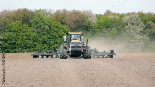 Tractor Preparing Field for Planting, A large tractor equipped with farming machinery plowing a vast field surrounded by dense green forest photo