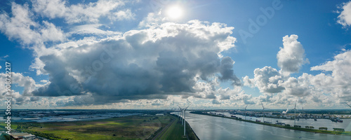 Massive cloud formations are present above the entrance of the Port of Rotterdam with greenhouses of the Westland region on the left, wind turbines in the middle, and oil refineries on the right.