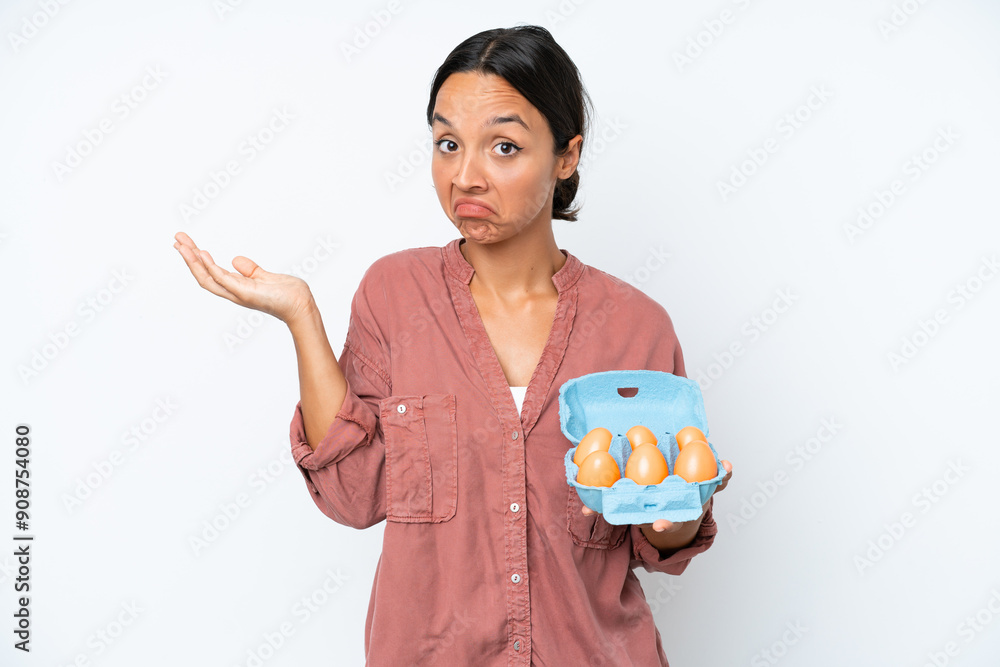 Young hispanic woman holding eggs isolated on white background having doubts while raising hands Young hispanic woman holding eggs isolated on white background having doubts while raising hands
