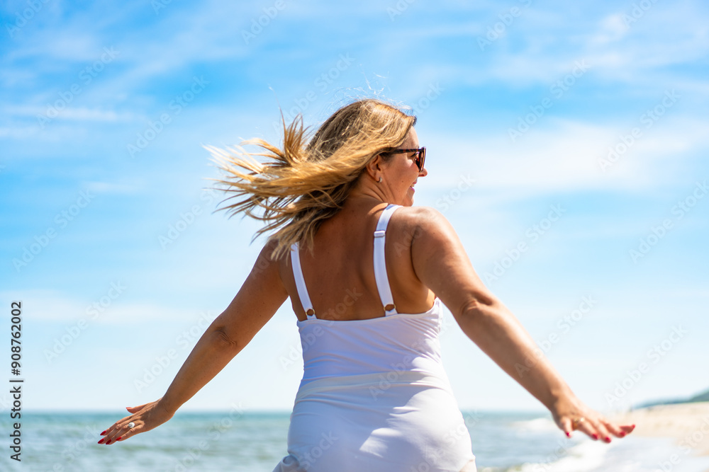 Naklejka premium Summer vacation by Baltic Sea. Beautiful elegant woman in white swimsuit and white sarong with her arms spread out walking by sea on sandy beach on beautiful sunny day. Back view