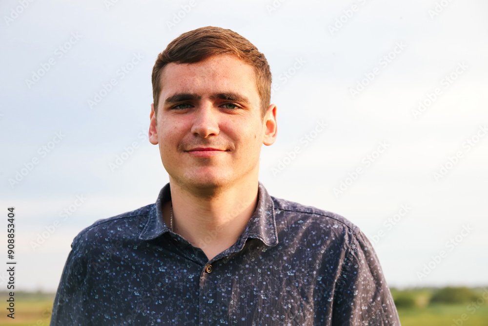 Young man smiling outdoors. Portrait of a 20s Caucasian male. Cheerful young man with brown hair wearing blue shirt against the sky Young man smiling outdoors. Portrait of a 20s Caucasian male. Cheerful young man with brown hair wearing blue shirt against the sky