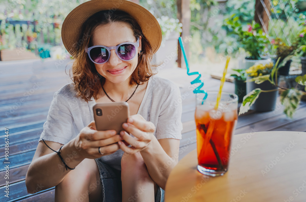Young cheerful woman in hat with refreshing cocktail with ice and smartphone in hands on summer terrace of cottage