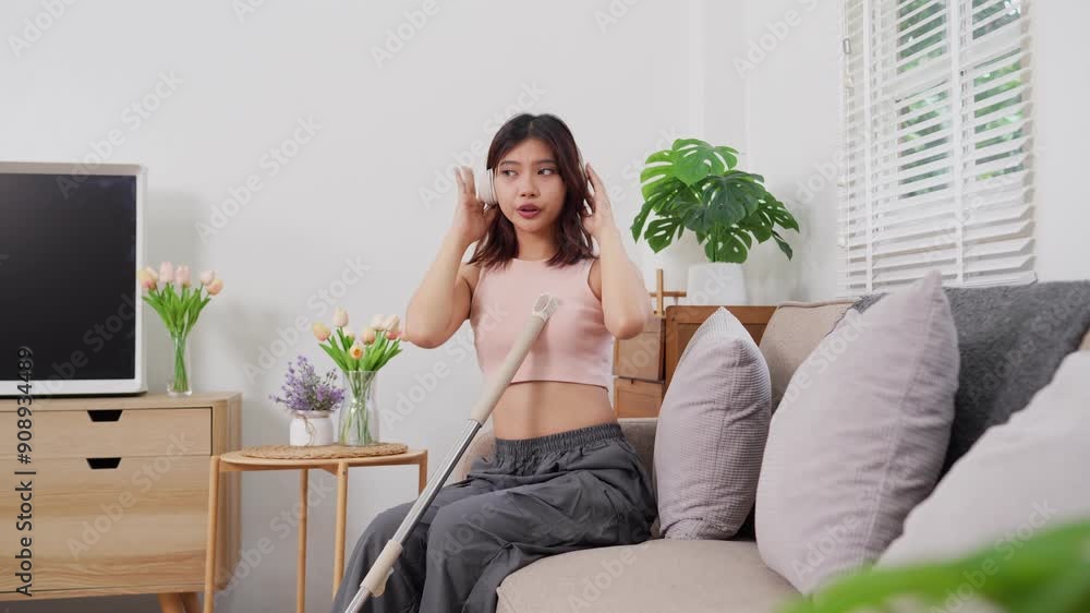 An Asian woman in casual clothing is dancing and singing into a mop in her living room, appearing cheerful and lively An Asian woman in casual clothing is dancing and singing into a mop in her living room, appearing cheerful and lively