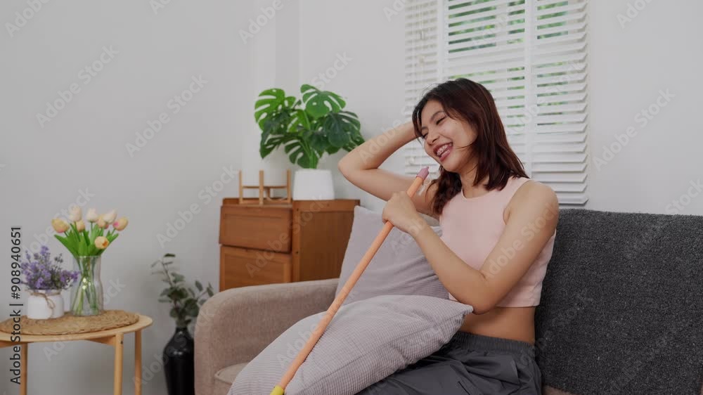 An Asian woman in casual attire is joyfully singing and dancing in her living room, holding a broom as a microphone, with a bright smile on her face An Asian woman in casual attire is joyfully singing and dancing in her living room, holding a broom as a microphone, with a bright smile on her face