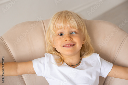 Little happy smiling boy at home with strabismus eyes, squinting eyes, cross-eyed. Child boy wearing white t-shirt on a beige chair near white wall