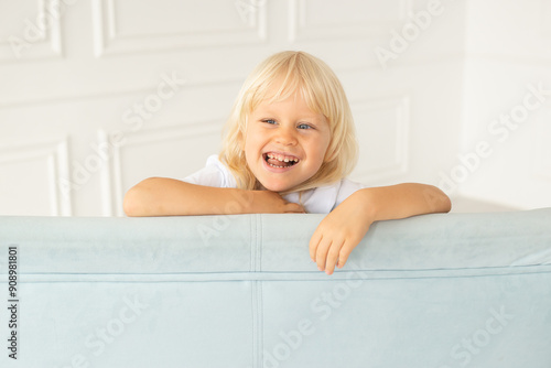 Laughing boy at home with strabismus eyes, squinting eyes. Child boy wearing white t-shirt on a blue sofa near white wall