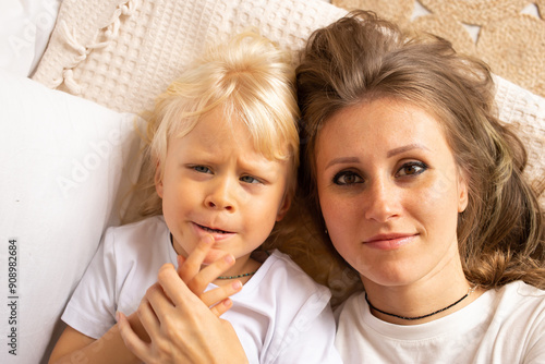 Mother and her little son lying on bed at home. Child boy with strabismus eyes, squinting eyes, cross-eye. Motherhood, affection