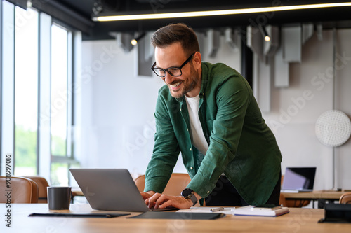 Young businessman in glasses smiling and typing over laptop computer while standing at desk in office