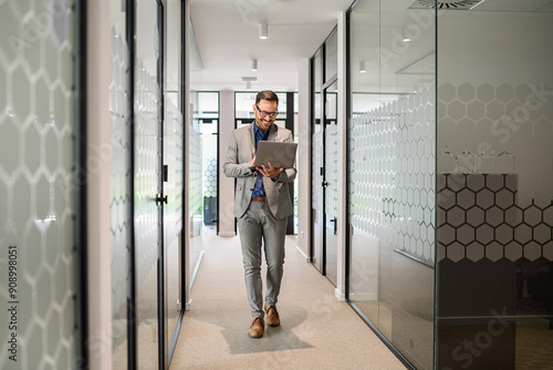 Full length of male software developer researching online over laptop while walking in office corridor