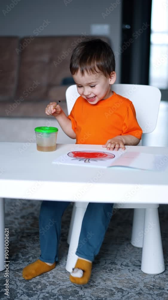 Cheerful kid sitting at the desk stepping his feet joyfully. Happy smiling toddler painting with a brush. Vertical video. Cheerful kid sitting at the desk stepping his feet joyfully. Happy smiling toddler painting with a brush. Vertical video.