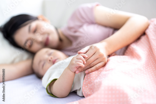 Asian woman lying with baby boy on the bed holding his little fingers. Happy baby boy was holding by his mother. Asian family with little boy at home enjoy tender family moment, motherhood.