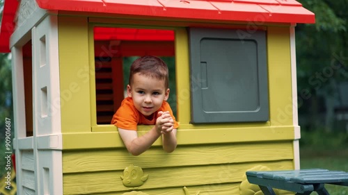 Lovely smiling toddler boy peeps out of the window in the playhouse. Happy child playing outdoors in summer at the playground. photo