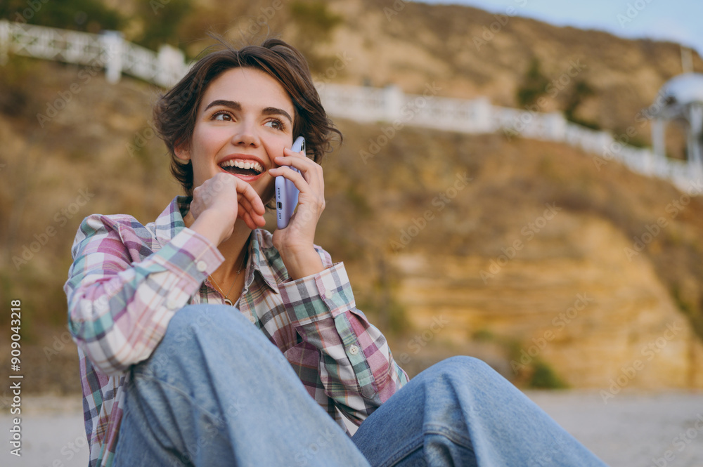 Young smiling happy cheerful woman she wearing shirt casual clothes talk speak on mobile cell phone sits rest on sea ocean sand shore beach outdoor seaside in summer day free time. Lifestyle concept. Young smiling happy cheerful woman she wearing shirt casual clothes talk speak on mobile cell phone sits rest on sea ocean sand shore beach outdoor seaside in summer day free time. Lifestyle concept.