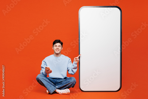 Full body young fun man wears blue sweatshirt casual clothes point on big huge blank screen mobile cell phone with area use smartphone isolated on plain red orange color background. Lifestyle concept. photo