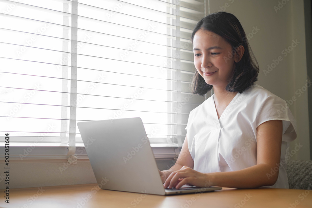 Young woman working on a laptop at home, sitting by a window with blinds. The image captures a productive remote work environment. Young woman working on a laptop at home, sitting by a window with blinds. The image captures a productive remote work environment.