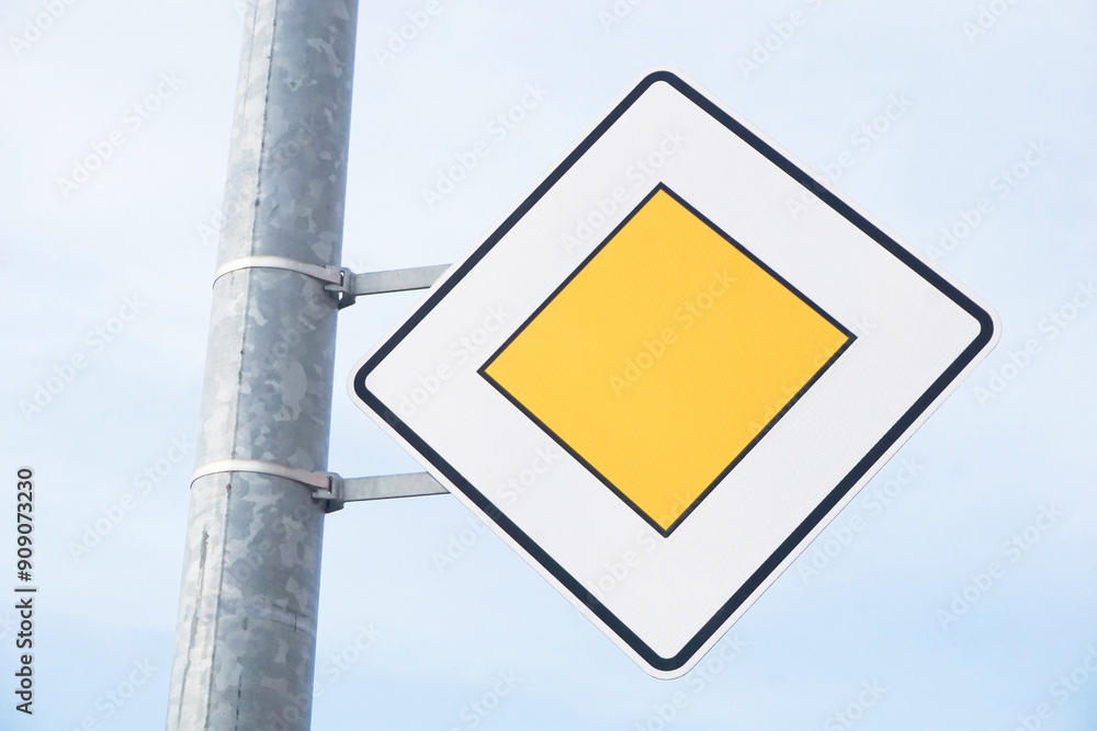Priority road sign. White and yellow diamond shape mounted on a metal pole. Traffic law texture. Indicating traffic regulations. Simple road rules background. Bright european road traffic. Priority road sign. White and yellow diamond shape mounted on a metal pole. Traffic law texture. Indicating traffic regulations. Simple road rules background. Bright european road traffic.