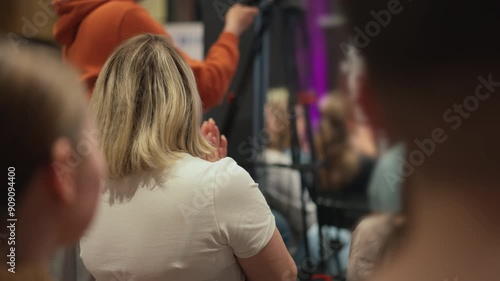 Woman with blond hair wearing a T-shirt applauds the speaker at presentation. Engaged crowd listens to presentation on latest innovations and technological breakthroughs at a tech conference.