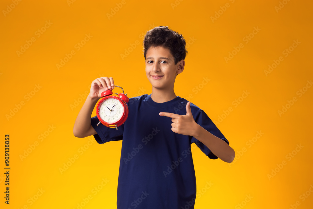 Child boy holding alarm clock and pointing with finger at it, over yellow background. Time management concept Child boy holding alarm clock and pointing with finger at it, over yellow background. Time management concept