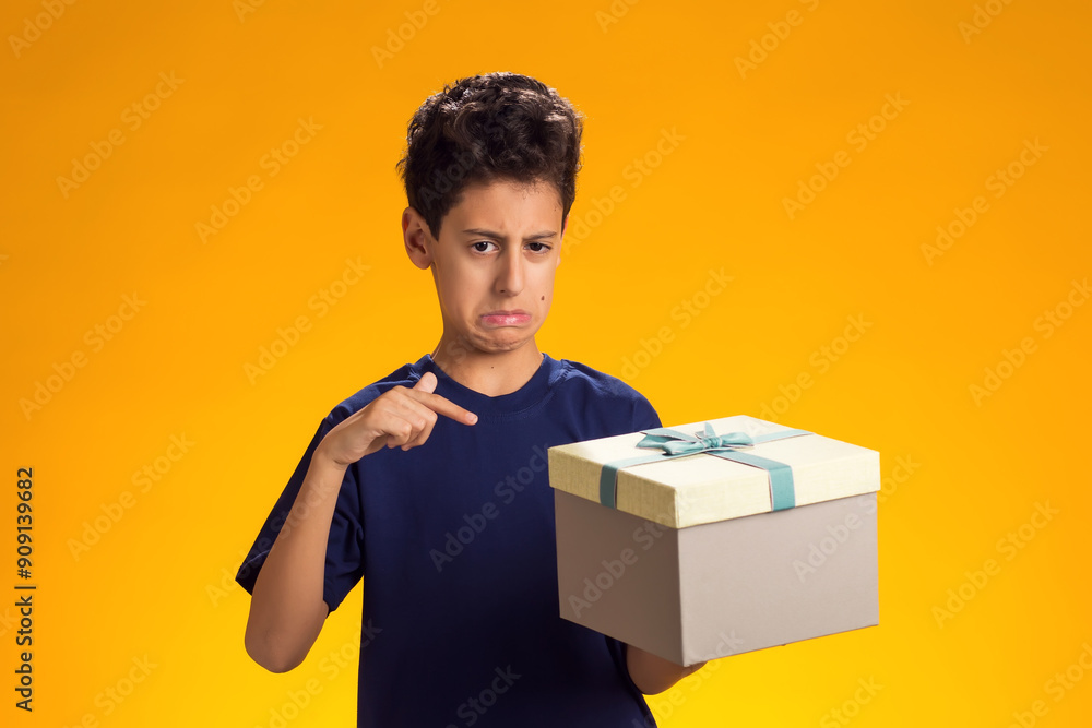 Angry child boy holding giftbox and pointing finger at it over yellow background. He doesn't like the gift. Birthday, celebration and disappointment concept Angry child boy holding giftbox and pointing finger at it over yellow background. He doesn't like the gift. Birthday, celebration and disappointment concept