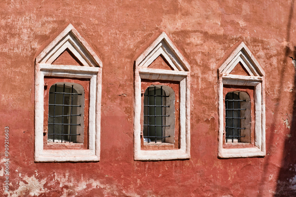Three windows with metal cage in a row on the wall of an old red building. Ancient stone building. Three windows with metal cage in a row on the wall of an old red building. Ancient stone building.