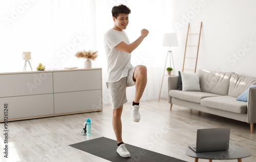 Online Workout Concept. Cheerful Smiling Asian Guy Training In Living Room, Doing Twist Knees To Elbow Exercise. Happy Man Standing And Lifting Leg Up, Using Laptop Watching Tutorial Lookign At Screen photo