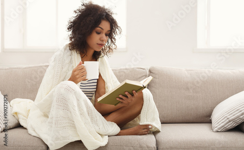 Thoughtful african-american student girl reading book. Young woman studying at home, sitting on beige couch wrapped up in white blanket and holding coffee cup, copy space photo