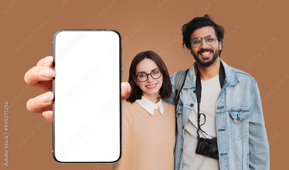 A young multiethnic couple pose together in front of a brown backdrop. They are holding a black smartphone with a blank white screen in front of them, both smiling and looking at the camera A young multiethnic couple pose together in front of a brown backdrop. They are holding a black smartphone with a blank white screen in front of them, both smiling and looking at the camera