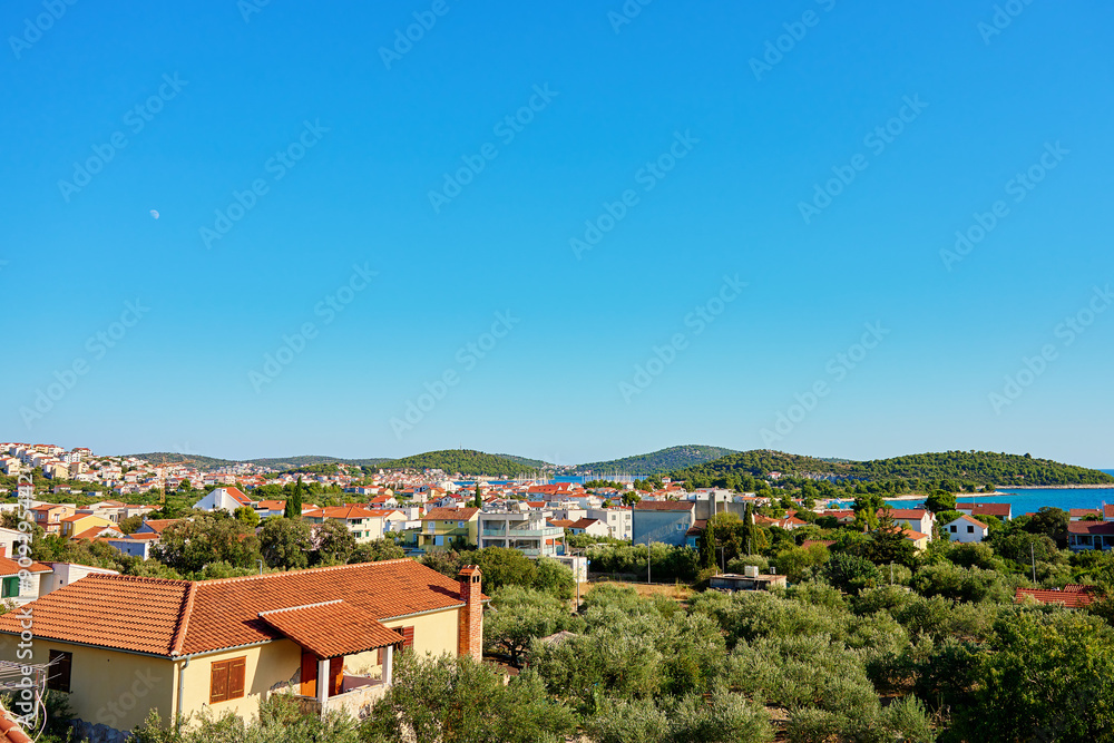 Obraz premium View on coastal town with red roofed houses and lush green hills under clear blue sky. Touristic Mediterranean town near Adriatic sea for summer holidays
