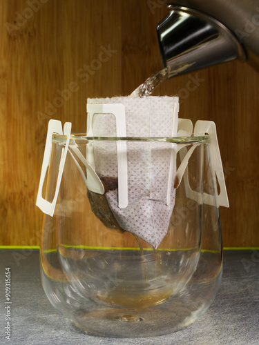 Closeup of boiling water from a kettle being poured over ground coffee in a filter bag, hanging over a glass cup by plastic ears, to brew.