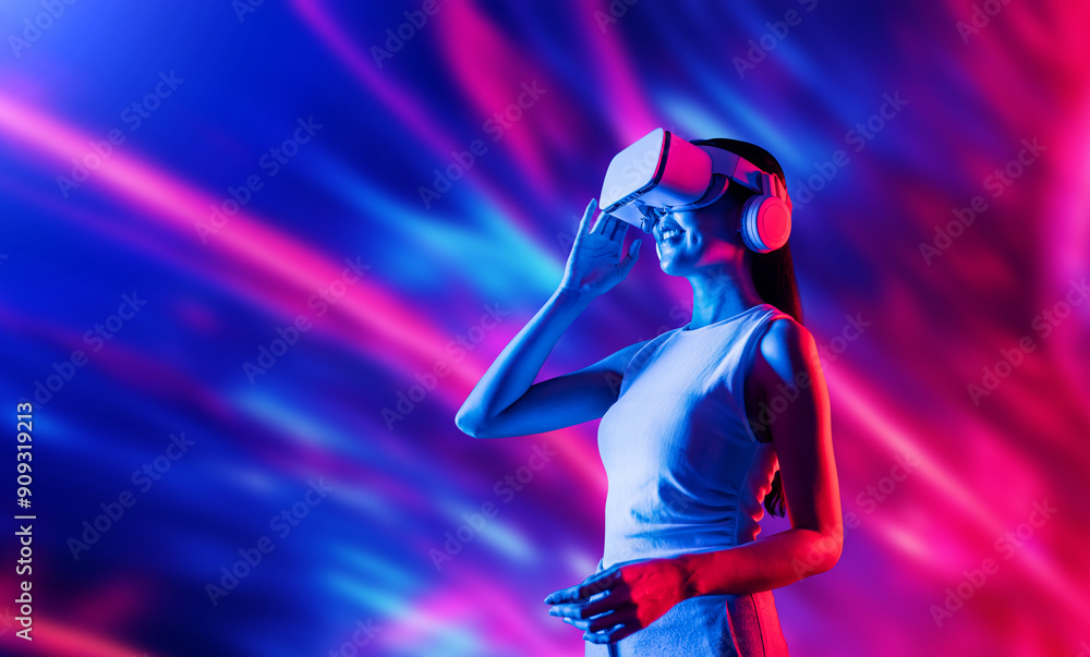 Smart female standing surrounded by neon light wearing VR headset connecting metaverse, future cyberspace community technology. Elegant woman looking faraway and smiling satisfactorily. Hallucination. Smart female standing surrounded by neon light wearing VR headset connecting metaverse, future cyberspace community technology. Elegant woman looking faraway and smiling satisfactorily. Hallucination.