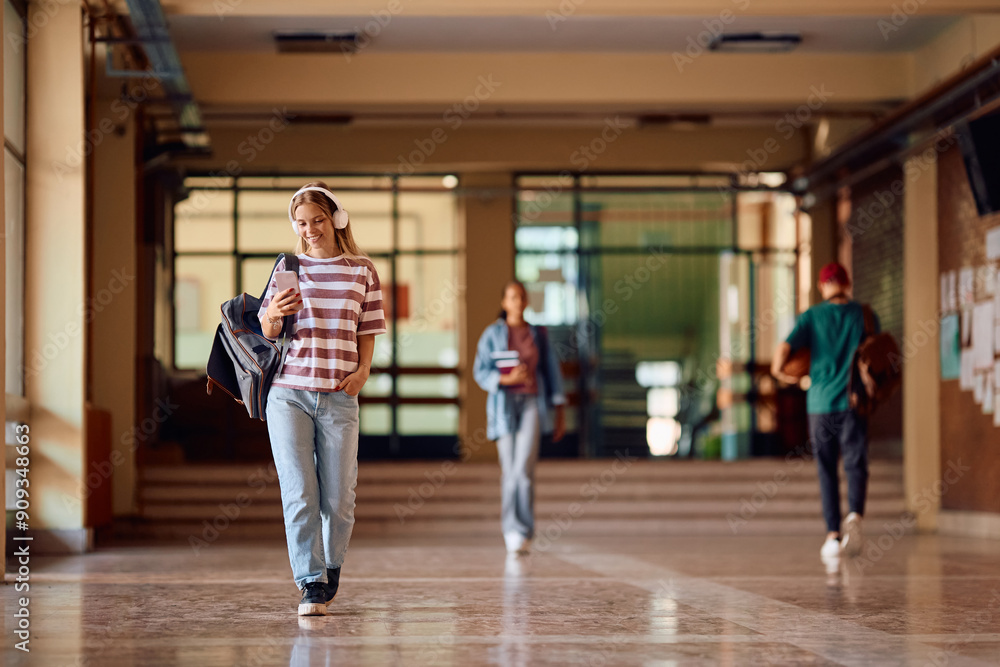 Obraz premium Happy teenage girl using mobile phone while walking through high school hallway.