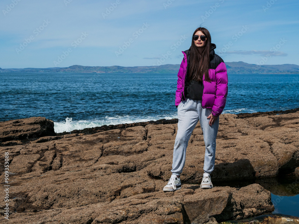 Φωτογραφία Teenager girl standing on rough stone coast, blue ocean and blue sky in the background