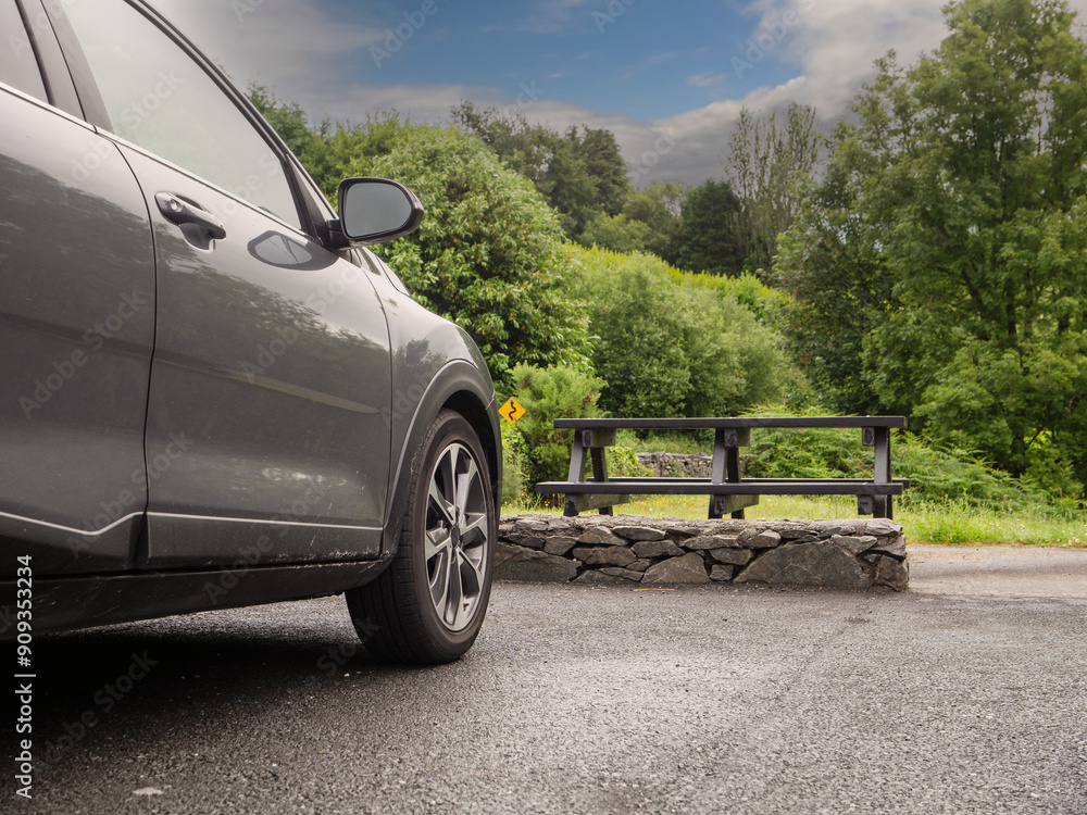 Φωτογραφία Black color car parked in a car park in a park close to a picnic area with table and bench, lush green forest in the background