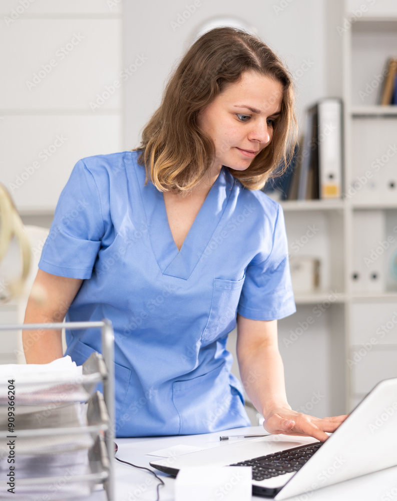 Φωτογραφία Portrait of female doctor in surgical scrubs standing at working table in her office and using laptop