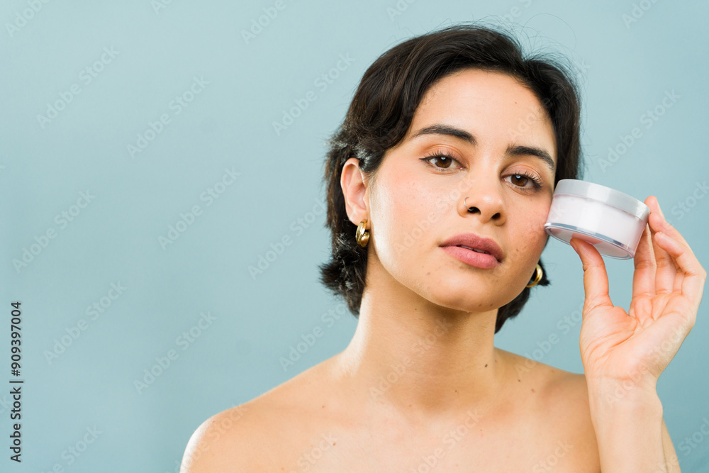 Serious-looking young woman holds a moisturizer jar near her face, presenting a beauty product Serious-looking young woman holds a moisturizer jar near her face, presenting a beauty product