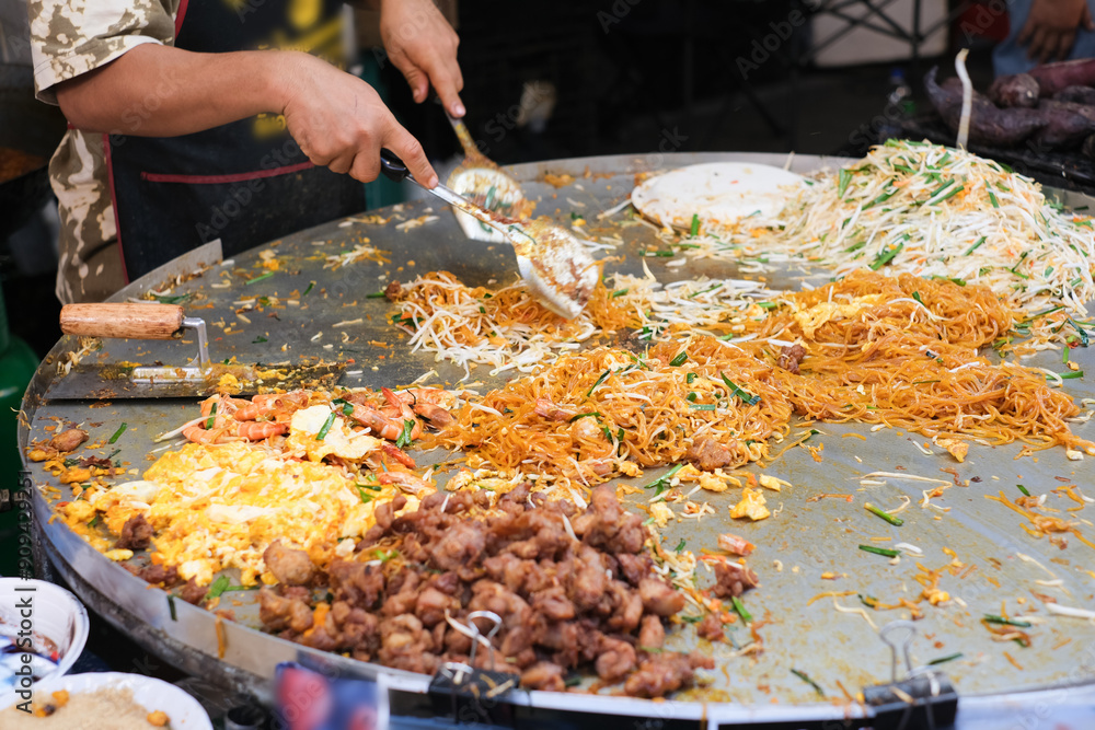 Obraz premium A man is cooking food on a large plate. The plate is full of different types of food, including noodles, meat, and vegetables. The man is using a spoon to serve the food onto plates