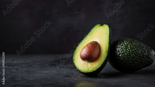 Close-up of fresh ripe avocados with one halved showing the seed, accompanied by green leaves on a dark textured background...