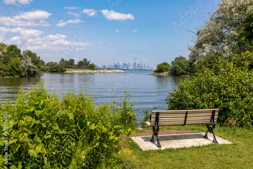 Canvas Print A wooden bench overlooks Lake Ontario at Sam Smith Park in Etobicoke, Ontario, framed by greenery, with a distant Toronto skyline on the horizon