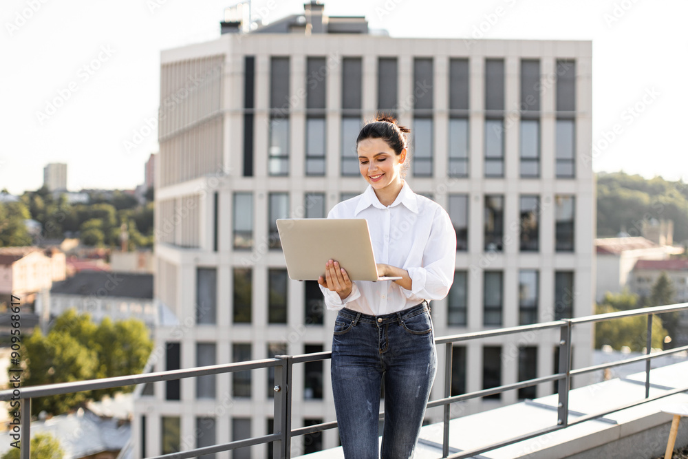Smiling young woman using laptop on rooftop with modern city skyline in background. Business professional enjoying outdoor workspace with urban scenery. Smiling young woman using laptop on rooftop with modern city skyline in background. Business professional enjoying outdoor workspace with urban scenery.