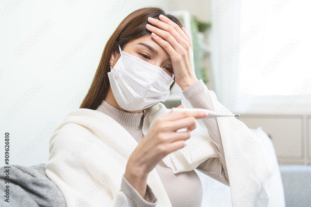 Sick, influenza asian young woman wearing face mask, have fever hand touching forehead, holding thermometer for check measure body temperature, illness while sitting rest on sofa at home. Health care. Sick, influenza asian young woman wearing face mask, have fever hand touching forehead, holding thermometer for check measure body temperature, illness while sitting rest on sofa at home. Health care.